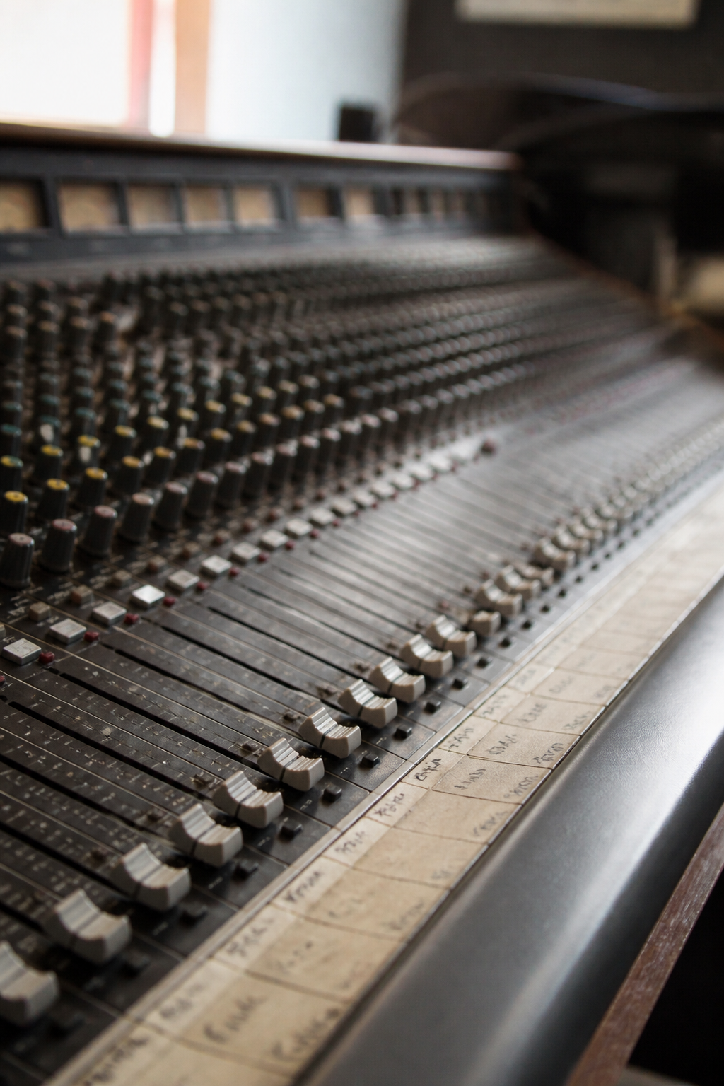A close view of a large analogue mixing desk, rows of faders and worn channel labels lit from one side