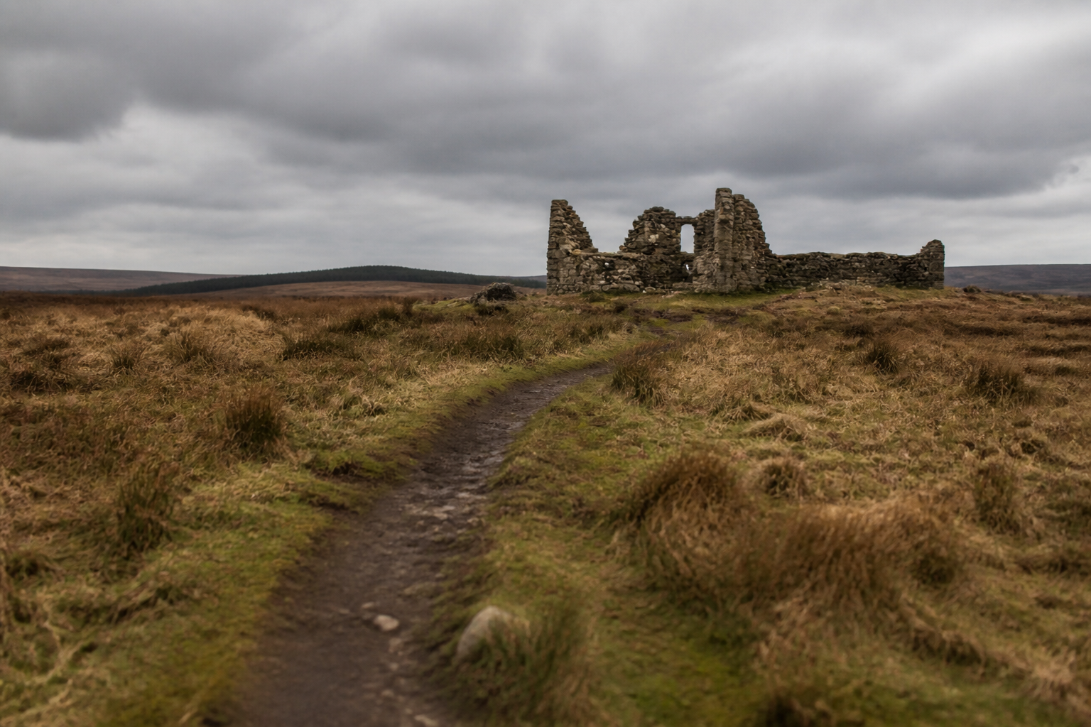 A worn footpath crossing open moorland towards a roofless stone ruin under a heavy grey sky
